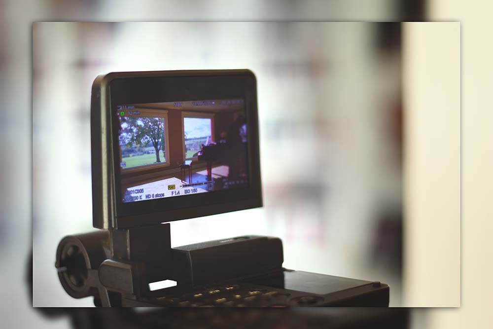 Photograph of a camera monitor depicting a big room with two large windows and a grand piano.
